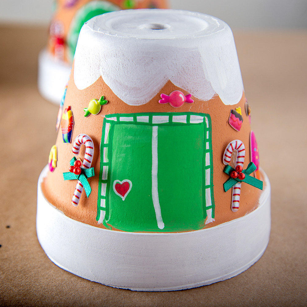 A flower pot is flipped upside down and painted to resemble a gingerbread house