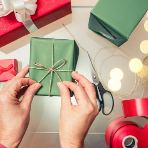 A person's hands tie a string around a green present. Various gift wrapping supplies are covering the table