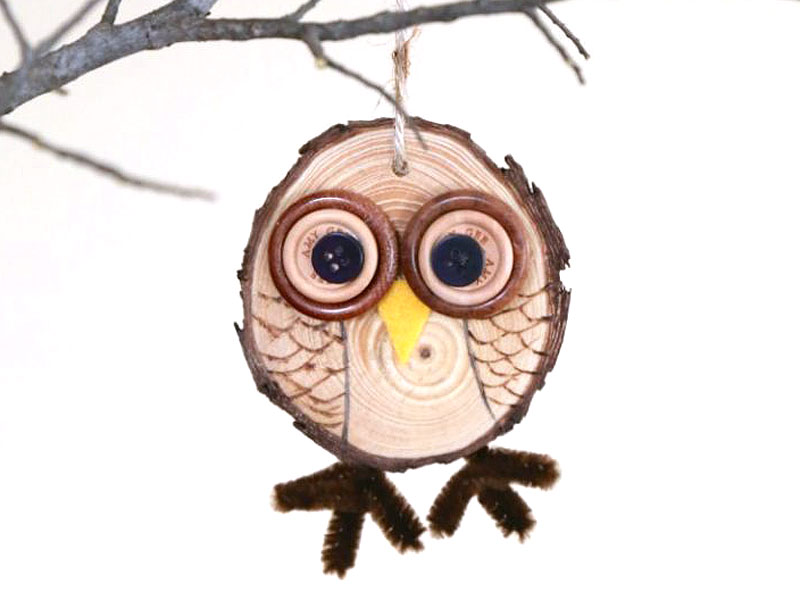 An owl ornament made out of a wood slice and various craft supplies hangs from a tree branch