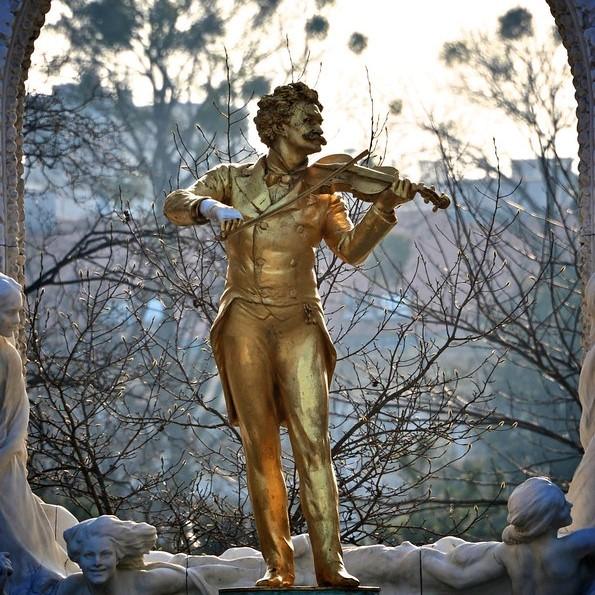 A bronze statue of Johann Strauss II playing the violin. The statue is outdoors, framed by a decorative stone arch. Tree branches and the Vienna skyline are visible in the background.